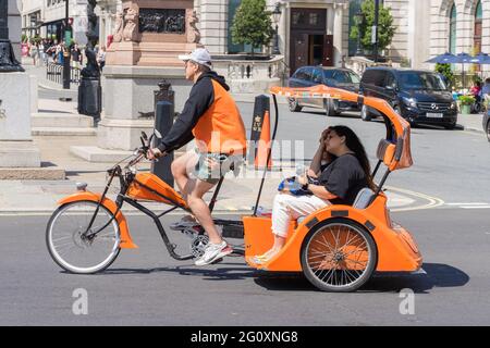 Eine orangefarbene Rikscha mit Touristen im Hintergrund, die an einem heißen Sommertag durch die Straßen Londons reiten. Stockfoto