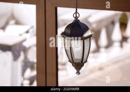 Eiserne Retro-Laterne mit Straßenbeleuchtung, die mit einer Glühbirne auf dem Hintergrund eines Fensters mit einem braunen Rahmen und Reflexionen im Glas aufgehängt ist Stockfoto