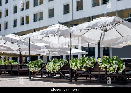 Weißer Sonnenschirm über den Tischen mit Sitzgelegenheiten auf der Terrasse eines Straßencafés, das an einem sonnigen Sommertag mit blühenden Petunienblumen geschmückt ist, niemand. Stockfoto
