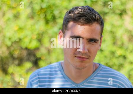 Defokussiertes Nahaufnahme traurig ernst junge Brünette kaukasischen Mann mit kurzen Haaren. Sommer verschwommen Natur Bokeh Hintergrund. Porträt eines hübschen jungen Mannes. O Stockfoto