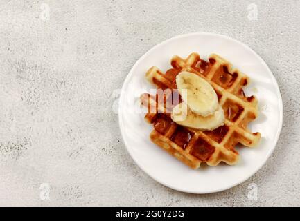 Wiener Waffel mit Bananenscheibe und Honig auf einem weißen runden Teller auf hellgrauem Hintergrund. Draufsicht, flach liegend Stockfoto