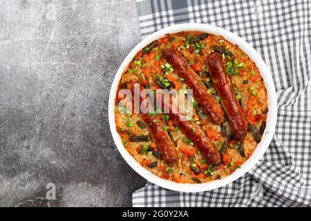 Gebackene Würstchen mit Reis und Gemüse in einer weißen Backform auf dunkelgrauem Hintergrund. Draufsicht, flach liegend Stockfoto