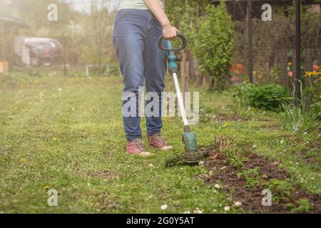 Frau mäht das Gras mit einem Rasentrimmer. Hobby und Gartenarbeit. Hof und Verbesserung. Stockfoto