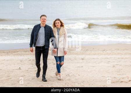 Attraktives junges Paar, das am Ufer eines Sandstrandes entlang geht, auf einem romantischen Frühlingsurlaub, im Freien.Paar, das einen Spaziergang macht und die Hände auf der Ba hält Stockfoto