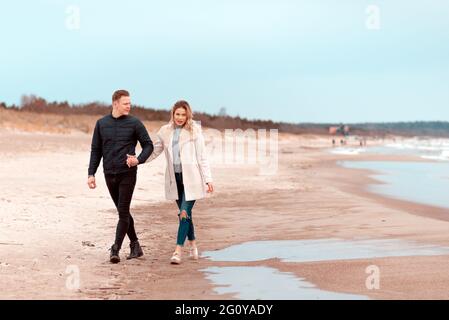 Attraktives junges Paar, das am Ufer eines Sandstrandes entlang geht, auf einem romantischen Frühlingsurlaub, im Freien.Paar, das einen Spaziergang macht und die Hände auf der Ba hält Stockfoto