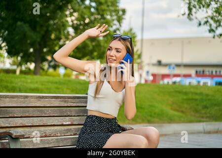 Glückliches Mädchen, das auf dem Handy anruft und auf einer Bank auf der Straße sitzt. Junge blonde Mädchen winkt zu einer Person in einem Park. Hochwertige Fotos Stockfoto