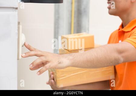 Asiatischer Mann mit Paket das Kontrollkästchen, um Klingeln kunden Klingel. Lieferung Mann in Orange einheitliche Läuten der Glocke. Home delivery Kurier Service und Versand conc Stockfoto