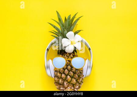 Ananas mit Sonnenbrille und kabellosen Kopfhörern mit weißer Frangipani-Blume auf gelbem Hintergrund. Minimales Sommerkonzept. Stockfoto