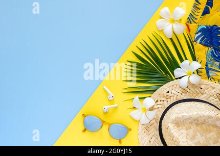 Sommer-Hintergrundkonzept mit Strandhut, Sonnenbrille, Kokosnussblättern, kabellosem Kopfhörer, Frangipani-Blumen und buntem Hemd auf gelbem und blauem Rücken Stockfoto