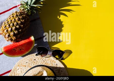 Sommer Hintergrund Konzept mit Sonnenbrille, Strandhut, Scheibe Wassermelone und Ananas auf Strandtuch mit gelbem Hintergrund und Schatten von Sonnenlicht. Stockfoto