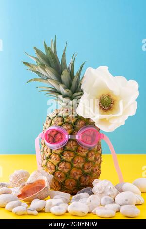 Ananas in rosa Schwimmbrille mit Seepsteinchen und Muscheln aus der Nähe auf gelb-blauem Hintergrund, wunderschöne weiße Blume, Sommerkonzept Stockfoto