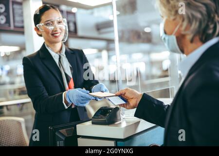 Der Check-in-Mitarbeiter der Fluggesellschaft trägt Schutzhandschuhe und einen Gesichtsschutz, der dem Passagier die Tickets aushändigt. Geschäftsreisende, die am Flughafen einchecken Stockfoto