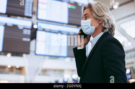 Mann mit Schutzmaske, der am Flughafen am Handy telefoniert. Ein reifer Geschäftsmann telefoniert am Flughafenterminal. Flugreisen während des Covid-19 Stockfoto