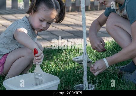 Kleine Mädchen und Jungen mit Bürsten putzen die Basis des jungen Baumstamms im Garten Stockfoto