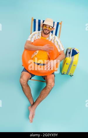 Blick aus der Höhe auf einen glücklichen Mann, der einen aufblasbaren Ring in der Nähe des Koffers hält und Pässe auf blauem Hintergrund Stockfoto