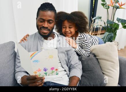 Vatertag. Afroamerikanische Tochter gibt dem Vater eine Grußkarte, die sie selbst gezeichnet hat und umarmt ihn zu Hause Stockfoto