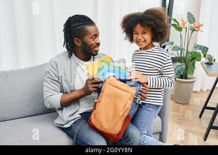 Vatertag. Vater hilft Tochter, farbige Copybooks vor ihrem ersten Schultag in die Schultasche zu legen. Glückliche Vaterschaft Stockfoto