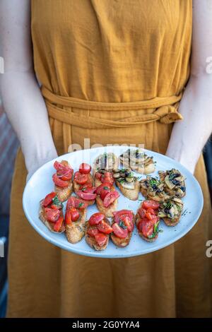 Eine Frau in einem orangefarbenen Kleid hält einen Teller mit hausgemachten Bruchetta mit Tomaten und Pilzen, Sommerfutter, vertikal Stockfoto