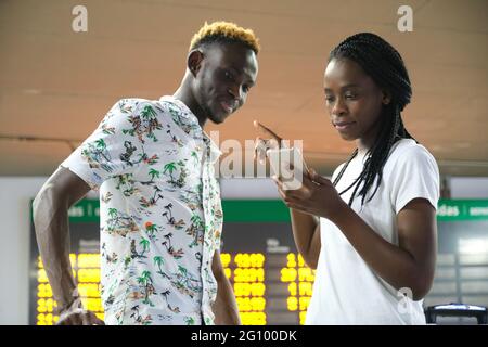 Junges afrikanisches Paar im Flughafen, das Koffer trägt, auf sein Smartphone schaut und mit Fluginformationen auf das Reisebüro zeigt. Stockfoto
