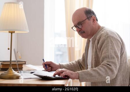 Leitender Mann, der mit Papierkram sitzt und den Rechner benutzt, während er Geld zählt Stockfoto