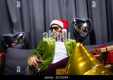 Junger arabischer Mann, der auf der Couch im Weihnachtsmütze sitzt und ein Glas Champagner auf grauem Hintergrund hält Stockfoto