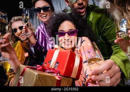 afroamerikanische Frau in Sonnenbrillen hält Geschenkboxen unter interracial Freunde in der Nähe grauen Vorhang auf schwarzem Hintergrund Stockfoto