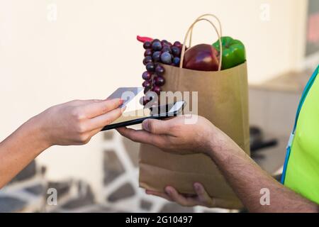 Erwachsene Hände mit Kreditkarte und Papiertüte. Beschnittene Nahaufnahme der weiblichen Kundin, die am Zahlungsterminal mit Kreditkarte bezahlt, Mann in gelber Unifo Stockfoto