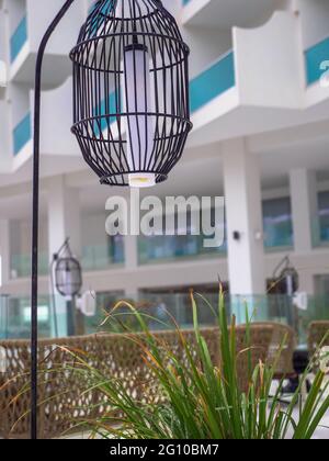 Hohe Laterne aus schwarzem Metallgitter, die in der Nähe der leeren Rattan-Wicket-braunen Stühle hängt, ohne dass nach dem Regen auf einer leeren Hotelterrasse Menschen stehen. Stockfoto