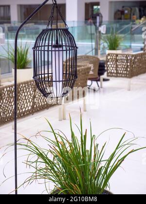 Hohe Laterne aus schwarzem Metallgitter, die in der Nähe der leeren Rattan-Wicket-braunen Stühle hängt, ohne dass nach dem Regen auf einer leeren Hotelterrasse Menschen stehen. Stockfoto