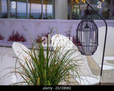 Hohe Laterne aus schwarzem Metallgitter, die an einer Kette in der Nähe leerer Rattan-Wicket-braune Stühle hängt, ohne dass nach dem Regen auf einer leeren Hotelterrasse Menschen stehen. Stockfoto