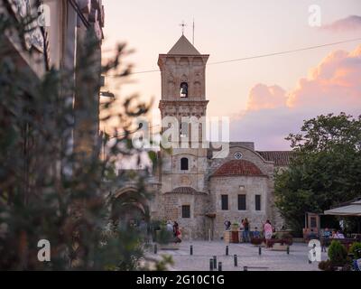 Blick durch Baumzweige über die st. Lazarus Kirche und Touristen sitzen und gehen auf dem Platz in der Nähe in Larnaca, Zypern. Historisches Erbe. Stockfoto