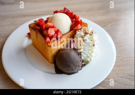 Honigtoast mit in Scheiben geschnittenen Erdbeeren, Vanille, dunklem Schokoladeneis und Schlagsahne auf weißem Teller Stockfoto