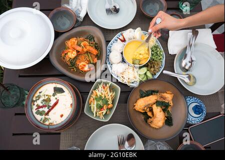 Draufsicht auf thailändisches Essen mit gesalzener Krabbe, Reisnudeln, gebratenem Seebarsch, gewürzter Mango, cremiger Hühnersuppe, Vermicelli Salat und Geschirr auf der TA Stockfoto