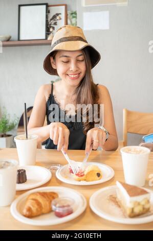 Glückliche junge asiatische Frau, die im Café auf einem Holztisch Dessert mit Croissant, Kuchen und Eiskaffee isst Stockfoto