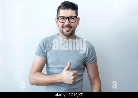 Ein glücklicher junger Kaukasusmann in einem grauen T-Shirt macht ein Daumenschild, das seine Unterstützung, seinen Respekt und seine Empfehlung für jemanden zeigt. Sprache des Körpers. I l Stockfoto