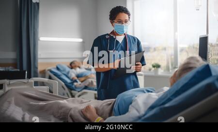 Krankenstation: Freundliche Krankenschwester trägt Gesichtsmaske spricht mit einer schönen älteren Patientin, die sich im Bett ausruht, überprüft ihre Vitalfunktionen, schreibt Informationen auf Stockfoto