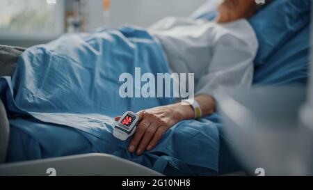 Krankenstation: Ältere Frau, die in einem Bett ruht, mit Finger-Herzfrequenzmonitor Pulsoximeter, der Puls zeigt. Ihre zerbrechlichen Hände ruhen auf einer Decke Stockfoto