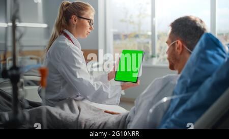 Krankenhausabteilung: Freundliche Ärztin spricht mit kranken männlichen Patienten, die im Bett ruhen, zeigt den Green Chroma Key Screen Tablet-Computer, erklärt die Ergebnisse Stockfoto