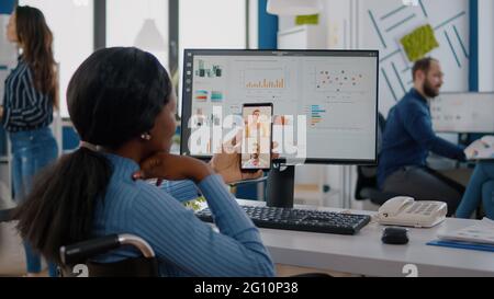 Rückansicht einer afrikanerin im Rollstuhl, die mit Freunden während eines Videoanrufs über Kopfhörer ein Smartphone hält und im Büro eine Pause einnimmt. Verschiedene Mitarbeiter planen Finanzprojekte Stockfoto