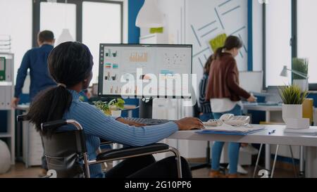 Junge afrikanische Arbeiterin mit Behinderungen, die statistische Informationen analysiert, die im Rollstuhl vor einem Finanzprojekt mit Computereingabe am Startup-Arbeitsplatz sitzen. Team prüft Unternehmensdaten Stockfoto