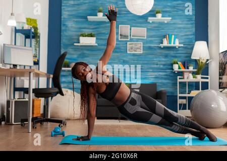 Fit athletische schwarze Frau, die in der Seitenplanke auf Yoga-Matte im Wohnzimmer zu Hause steht, Kraftübungen macht, für einen gesunden Lebensstil trainiert. Volle Länge der afroamerikanischen Arbeit. Stockfoto