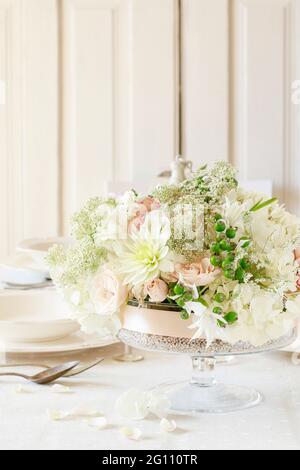 Blumenschmuck auf Sommerparty-Tisch. Kerze in Glasvase, Blumenkranz um sie herum. Weiße Teller, Tischtücher, romantischer Hochzeitsempfang. Stockfoto