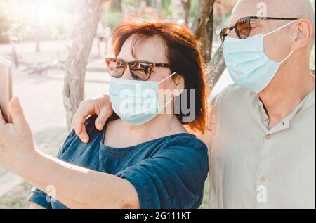 Rentnerpaar machen ein Selfie mit einer chirurgischen Maske - Rentner mittleren Alters machen während der Corona-Virus-Pandemie mit ihrem Smartphone ein Foto - warm Stockfoto