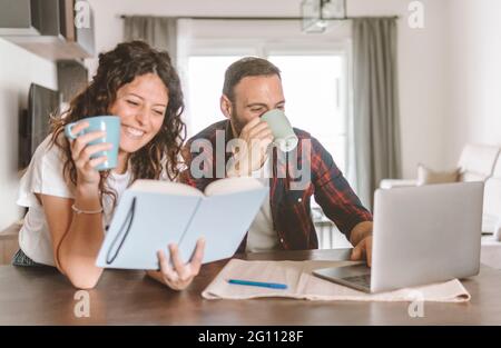 Ein verliebtes Paar, das zusammen mit einer Tasse Kaffee frühstückt - ein junges Paar trinkt eine Tasse Tee, während es den Laptop benutzt und ein Buch liest Stockfoto