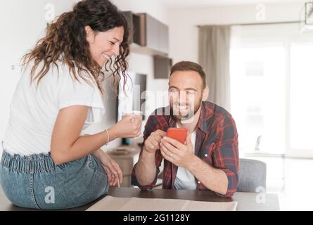 Das junge Paar lacht, während es sein Smartphone benutzt – das verliebte Paar frühstückt zusammen und liest die neuesten Nachrichten per Handy Stockfoto