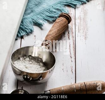 Messlöffel mit weißer Zutat vielleicht Mehl Zucker oder Puderzucker auf einem Tisch mit einem blauen Tuch und natürlichem Licht Stockfoto