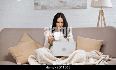 Kranke Frau, die mit einem Laptop sitzt und eine Tasse Tee hält Stockfoto