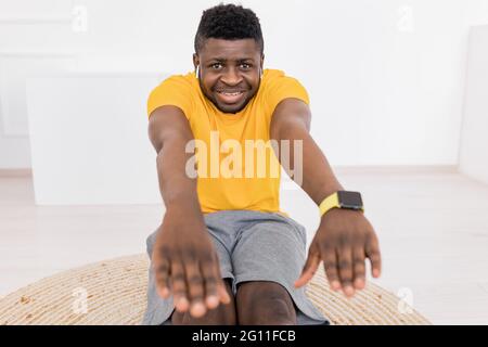 Sportlicher afroamerikanischer Mann, der Bein- und Rückenmuskeln vor der Kamera streckt, die Kamera anschaut, Musik hört und sich zu Hause aufwärmt Stockfoto