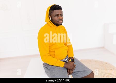Nachdenkliche afroamerikaner tragen einen Mantel und schauen auf die Kamera, während sie auf dem Tisch sitzen. Nachdenklicher afroamerikanischer Mann, der einen gelben Hoodie trägt und aussieht Stockfoto