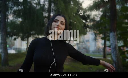 Lächelnde junge, schöne Frau mit Kopfhörern hört Musik und tanzt im Park in Zeitlupe Stockfoto